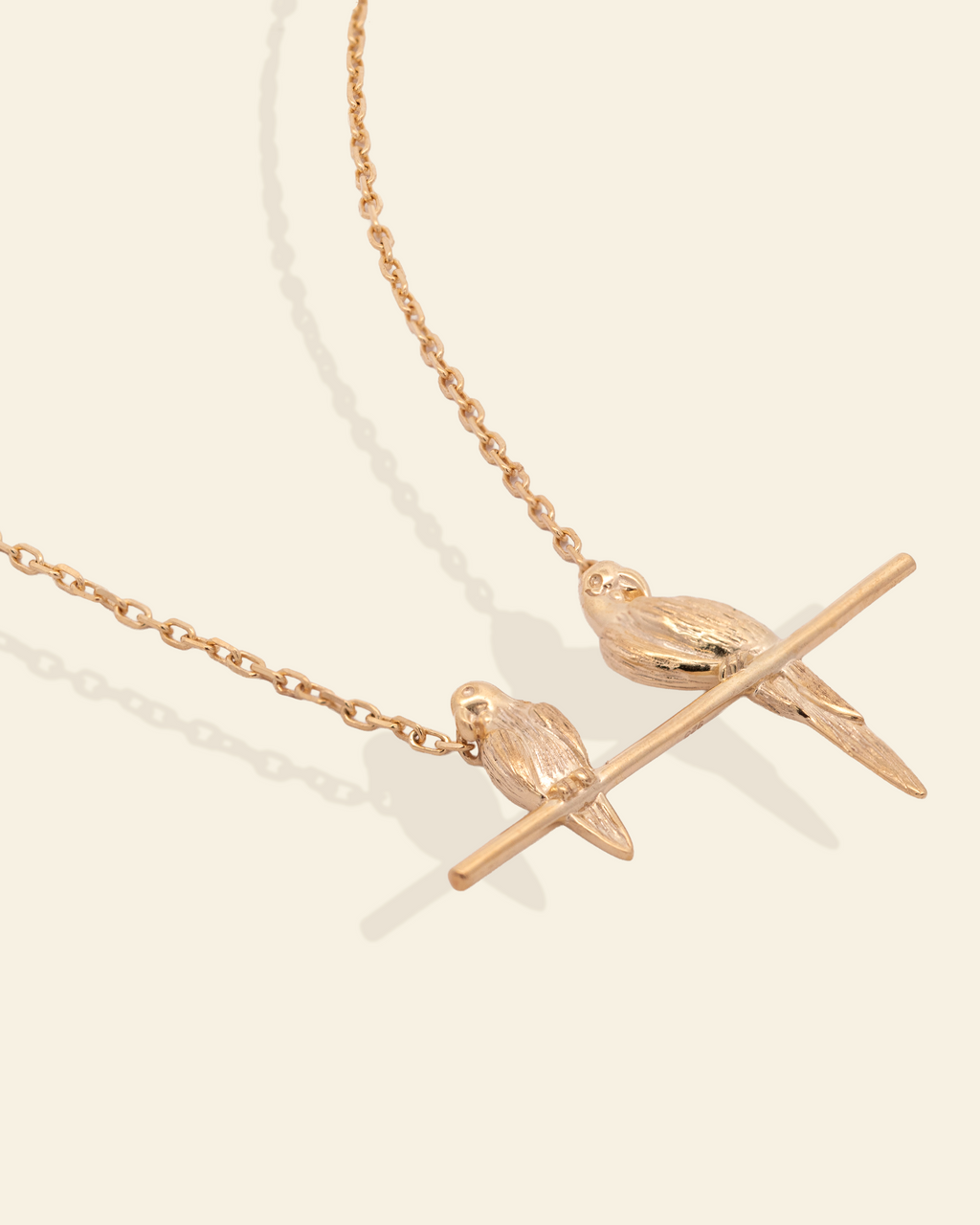 A delicate rose gold necklace featuring two small bird charms perched on a horizontal bar, displayed on a clean, light cream background.